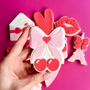 Puede incluir: Un conjunto de galletas de azúcar con temática de San Valentín decoradas con glaseado rosa, rojo y blanco. Las galletas tienen forma de corazón, lazo, labios, carta y la Torre Eiffel.
