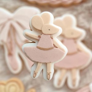 Peut inclure: Un biscuit en forme de souris ballerine rose et or. La souris porte un tutu rose et des chaussures dorées. Le biscuit est décoré d'une finition scintillante.