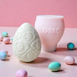 Könnte beinhalten: Ein weißes, aufwendig gemustertes Osterei steht neben einem hellrosa Behälter mit dem eingeprägten Schriftzug "DMC". Pastellfarbene Bonbon-Eier in Blau, Rosa und Grün sind um die Objekte herum auf einem rosa Hintergrund verstreut. Das Ei hat ein detailliertes Blumendesign.