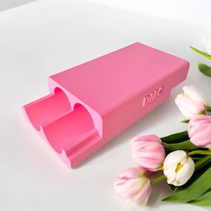 May include: A bright pink rectangular object with two cylindrical openings. The letters "DMG" are printed on the side. The object is on a white surface, with pink and white tulips in the background.