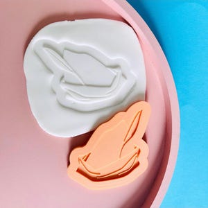 May include: A white silicone mold and a peach-colored cutter, both in the shape of a hat with a feather. The objects are on a pink tray with a blue background. The mold is used for creating decorative items.