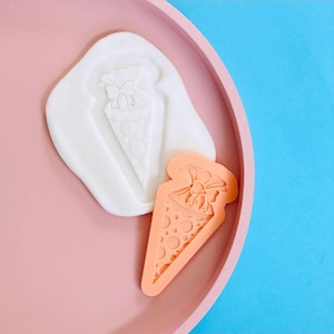 Peut inclure: Un moule en silicone blanc et un tampon en silicone orange, tous deux en forme de sac cadeau avec un nœud. Le sac cadeau a un motif à pois. Les articles sont sur un plateau rose avec un fond bleu.