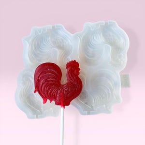 May include: A red rooster-shaped lollipop on a white stick, next to a white silicone mold with rooster designs. The mold has four rooster impressions. The background is a soft pink.
