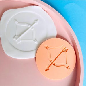 Könnte beinhalten: Zwei Wachssiegel mit einem Pfeil-und-Bogen-Design. Ein Siegel ist weiß, das andere pfirsichfarben. Beide Siegel sind rund und weisen das gleiche Design von Pfeil und Bogen auf. Die Siegel befinden sich auf einem rosa Tablett mit blauem Hintergrund.