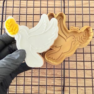 Könnte beinhalten: Ein weißer Keks in Taubenform mit einem gelben Olivenzweig, neben einem braunen Kunststoff-Ausstecher. Der Keks liegt auf einem schwarzen Drahtgitter. Der Ausstecher hat die Buchstaben "DMC".