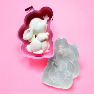 Peut inclure: Un moule en forme de lapin blanc avec une bordure rose, contenant une figurine de lapin blanc. Un moule transparent en forme de lapin est à côté. Les moules sont sur un fond rose. Le mot "DMC" est imprimé sur le moule rose.