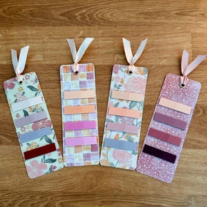 May include: Four pink and white patterned bookmarks with a ribbon attached to the top. Each bookmark has five horizontal stripes of different shades of pink, peach, and purple.