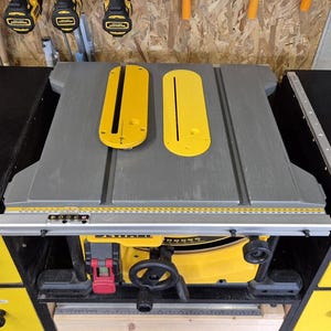 Może przedstawiać: Żółto-czarna piła stołowa DEWALT z szarym blatem i dwoma żółtymi wkładkami. Piła ma czarną podstawę z żółtymi szufladami i czerwonym wyłącznikiem bezpieczeństwa. Narzędzia wiszą w tle.