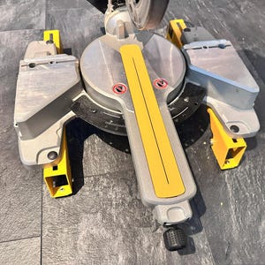 Può includere: Una troncatrice argentata e gialla con base nera. La sega ha un'impugnatura gialla e dispositivi di sicurezza. La lama è parzialmente visibile. La sega è progettata per tagliare legno e altri materiali.