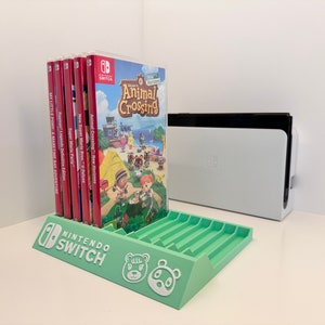 Peut inclure: Un support de cartouche de jeu Nintendo Switch en plastique vert avec des emplacements pour plusieurs jeux. Le support porte le logo Nintendo Switch et un personnage blanc d'Animal Crossing sur le devant. Le support est posé sur une surface blanche avec une console Nintendo Switch en arrière-plan.