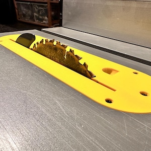 Può includere: Primo piano di una lama per sega da tavolo con un proteggi lama in plastica gialla. La lama è parzialmente visibile e ha una finitura argentata.