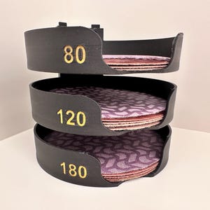 Puede incluir: Un organizador de discos de lija de plástico negro. El organizador tiene tres niveles, cada uno con un grano diferente: 80, 120 y 180. Cada nivel tiene una pila de discos de lija morados.