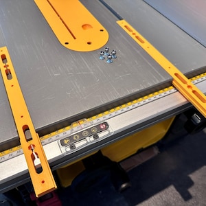 Op de afbeelding: Een close-up van een tafelzaag met twee gele hekverlengingen. De verlengingen hebben meerdere gaten en een liniaal die in centimeters is gemarkeerd. De tafelzaag heeft een doorzichtige plastic kap over de bedieningselementen.