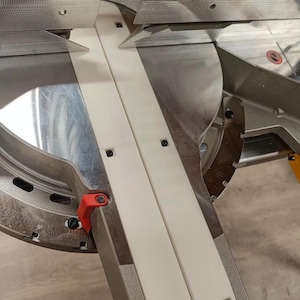 Op de afbeelding: Close-up van een witte kunststof verstekzaagbeugel met een zilveren metalen basis. De beugel heeft een groef in het midden en is vastgeschroefd. Het zaagblad is zichtbaar op de achtergrond.