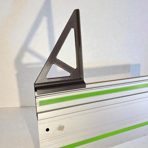 Peut inclure: Un équerre métallique gris foncé avec une découpe triangulaire est fixée à une glissière métallique argentée et verte. La glissière comporte deux bandes vertes vives. Ces outils sont probablement utilisés pour le travail du bois ou la construction.