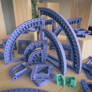Op de afbeelding: Een assortiment lichtpaarse plastic constructieonderdelen, waaronder gebogen en vierkante vormen, met kleine gaatjes. Er zijn ook twee kleine groene stukken aanwezig. De onderdelen zijn gerangschikt op een licht houten oppervlak, met houten blokken op de achtergrond.