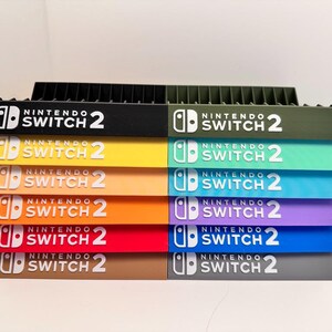 Peut inclure: Une pile de supports colorés pour console de jeux Nintendo Switch 2. Chaque support est d'une couleur différente, notamment noir, vert, jaune, orange, rouge, violet, bleu et marron. Le logo Nintendo Switch 2 est affiché en blanc.