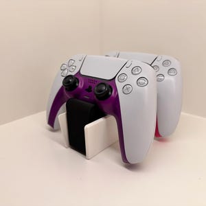 Pode incluir: Dois controladores PlayStation 5 brancos com detalhes roxos são exibidos em um suporte branco. Os controladores estão posicionados lado a lado, com a parte frontal dos controladores voltada para o observador.