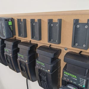 Könnte beinhalten: Ein hölzerner, an der Wand montierter Organizer mit mehreren schwarzen Batterieladegeräten und Halterungen. Die Ladegeräte sind mit der Marke "Festool" und Modellnummern beschriftet. Die Halterungen sind über den Ladegeräten montiert.
