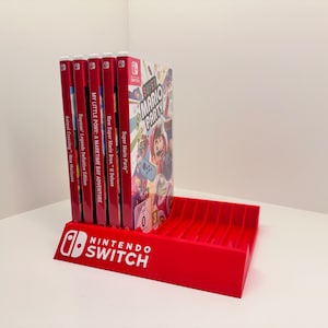 Puede incluir: Un soporte rojo para juegos de Nintendo Switch con el logotipo de Nintendo Switch en blanco. Contiene varias carcasas de juegos, incluyendo "Super Mario Party" y "Animal Crossing: New Horizons".
