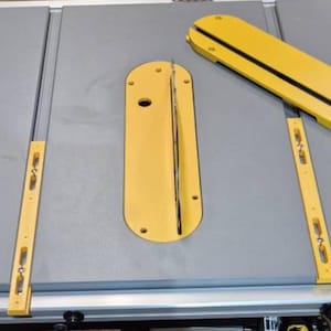 Peut inclure: Gros plan sur une scie à table avec deux inserts en plastique jaune. Les inserts ont une fente pour une lame et un trou pour une vis.