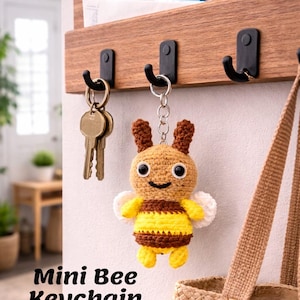 Puede incluir: Un llavero de abeja de ganchillo hecho a mano con una cara sonriente, rayas amarillas y marrones y alas blancas cuelga de un gancho negro. El llavero está junto a un juego de llaves y un organizador de pared de madera.