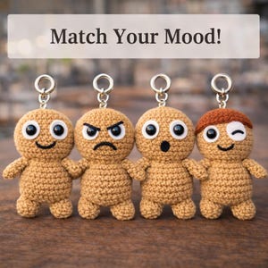 May include: Four crocheted keychains with different facial expressions: happy, angry, surprised, and winking. Each keychain is a tan color with black and white eyes and a silver keyring. The text "Match Your Mood!" is above the keychains.