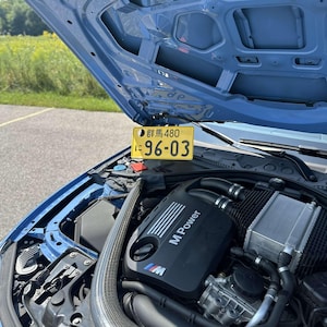 May include: A light blue car engine bay with the bonnet open. The engine is black with "M Power" printed on it. A yellow license plate with Japanese characters and the numbers "96-03" is visible.