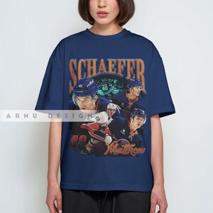 May include: Navy blue t-shirt featuring a vintage-style graphic of a hockey player with the name "SCHAEFFER" in orange lettering. The number "48" and the name "Matthew" are also visible. The shirt is worn by a person.
