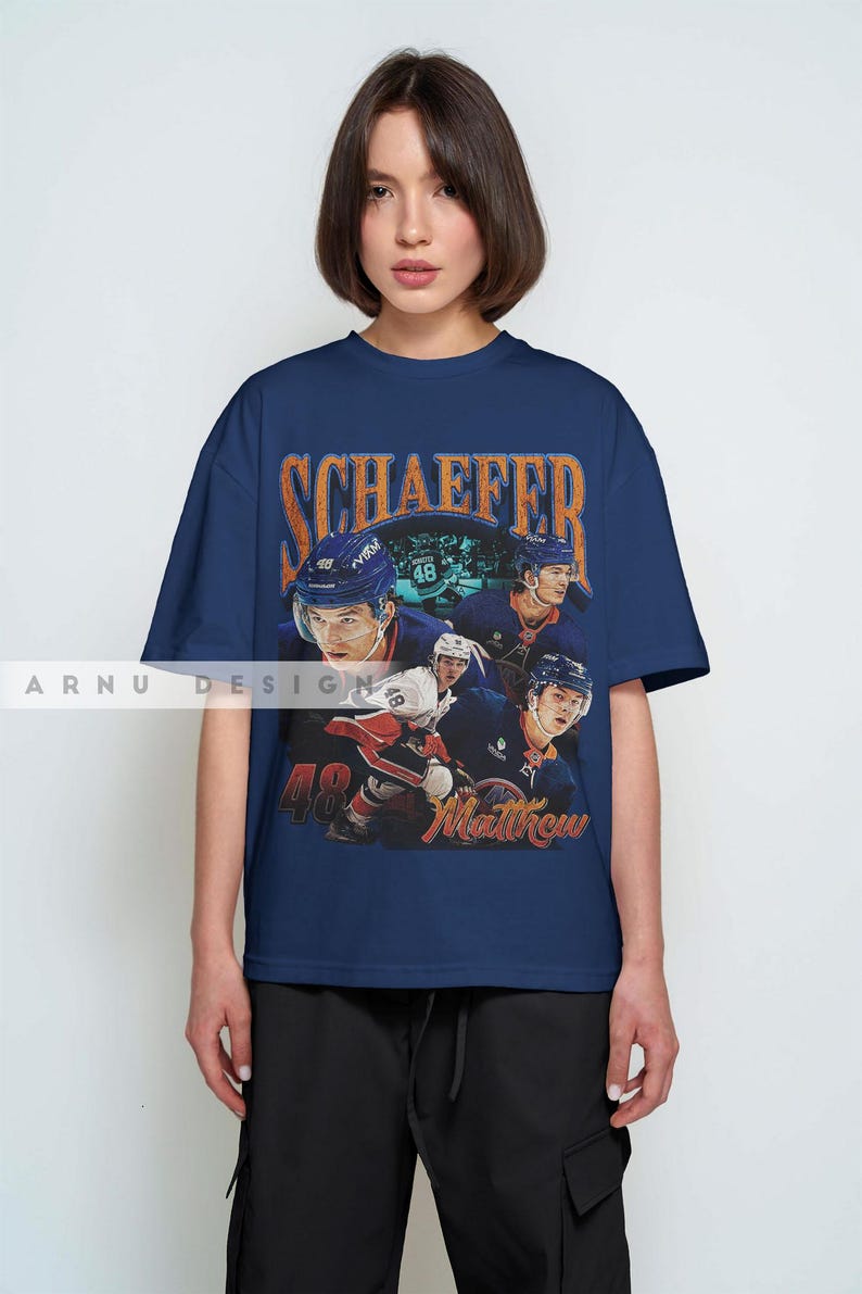 May include: Navy blue t-shirt featuring a vintage-style graphic of a hockey player with the name "SCHAEFFER" in orange lettering. The number "48" and the name "Matthew" are also visible. The shirt is worn by a person.
