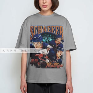 May include: A gray t-shirt featuring a vintage-style graphic of hockey players in blue and white uniforms. The shirt has the name "SCHAEFFER" and the number "48" printed on it, along with the name "Matthew". The shirt is from "ARNU DESIGN".