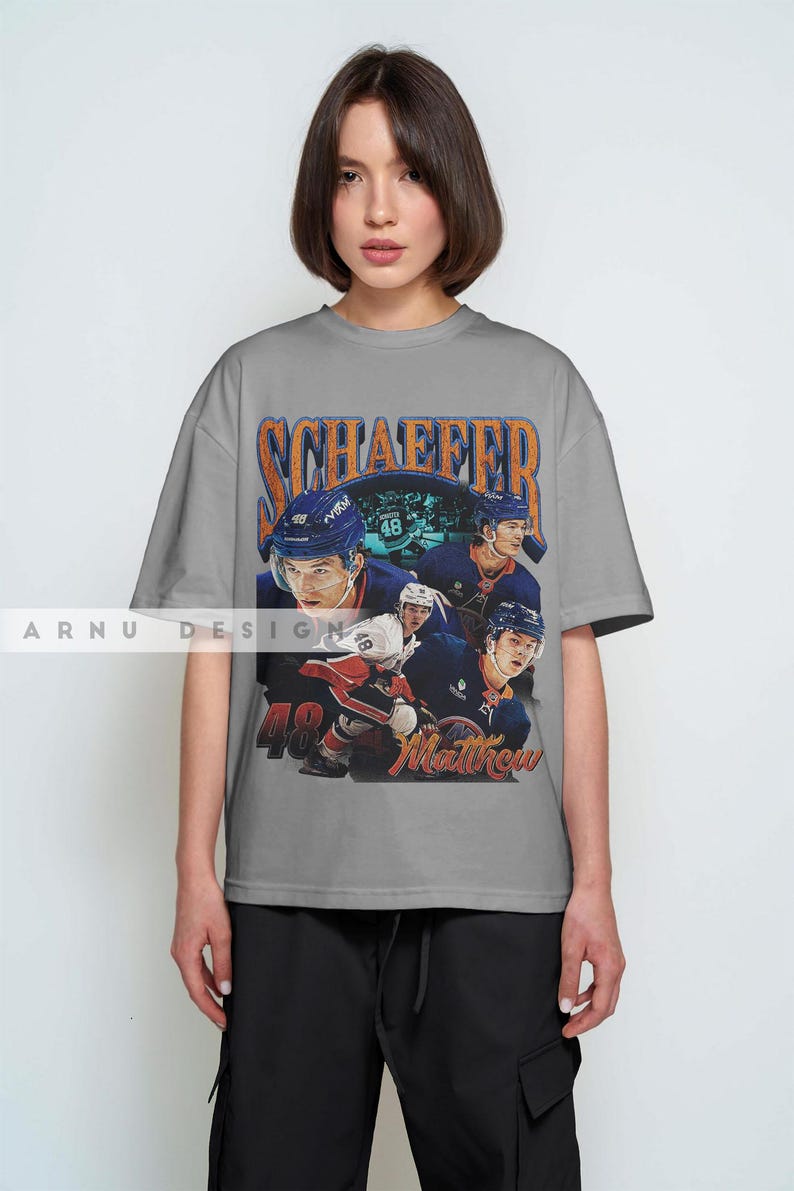 May include: A gray t-shirt featuring a vintage-style graphic of hockey players in blue and white uniforms. The shirt has the name "SCHAEFFER" and the number "48" printed on it, along with the name "Matthew". The shirt is from "ARNU DESIGN".