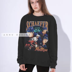May include: Black sweatshirt featuring a hockey player design. The design includes the name "SCHAEFFER" in orange and blue, the number "48", and the name "Matthew". The sweatshirt has a crew neck.