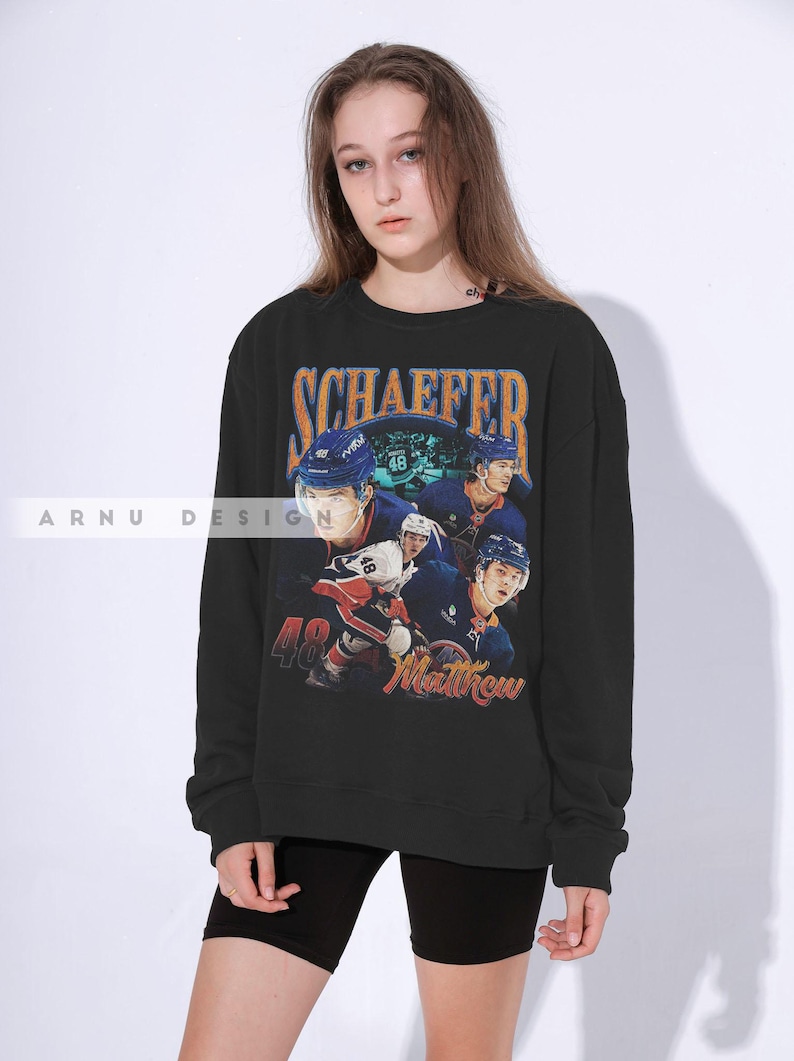 May include: Black sweatshirt featuring a hockey player design. The design includes the name "SCHAEFFER" in orange and blue, the number "48", and the name "Matthew". The sweatshirt has a crew neck.