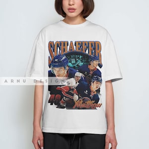 May include: White t-shirt featuring a vintage-style graphic of a hockey player with the name "SCHAEFFER" and the number "48". The design includes multiple images of the player in action. The word "Matthew" is also visible.