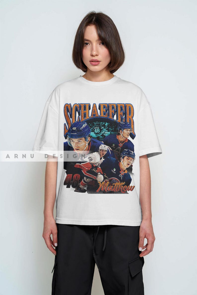May include: White t-shirt featuring a vintage-style graphic of a hockey player with the name "SCHAEFFER" and the number "48". The design includes multiple images of the player in action. The word "Matthew" is also visible.