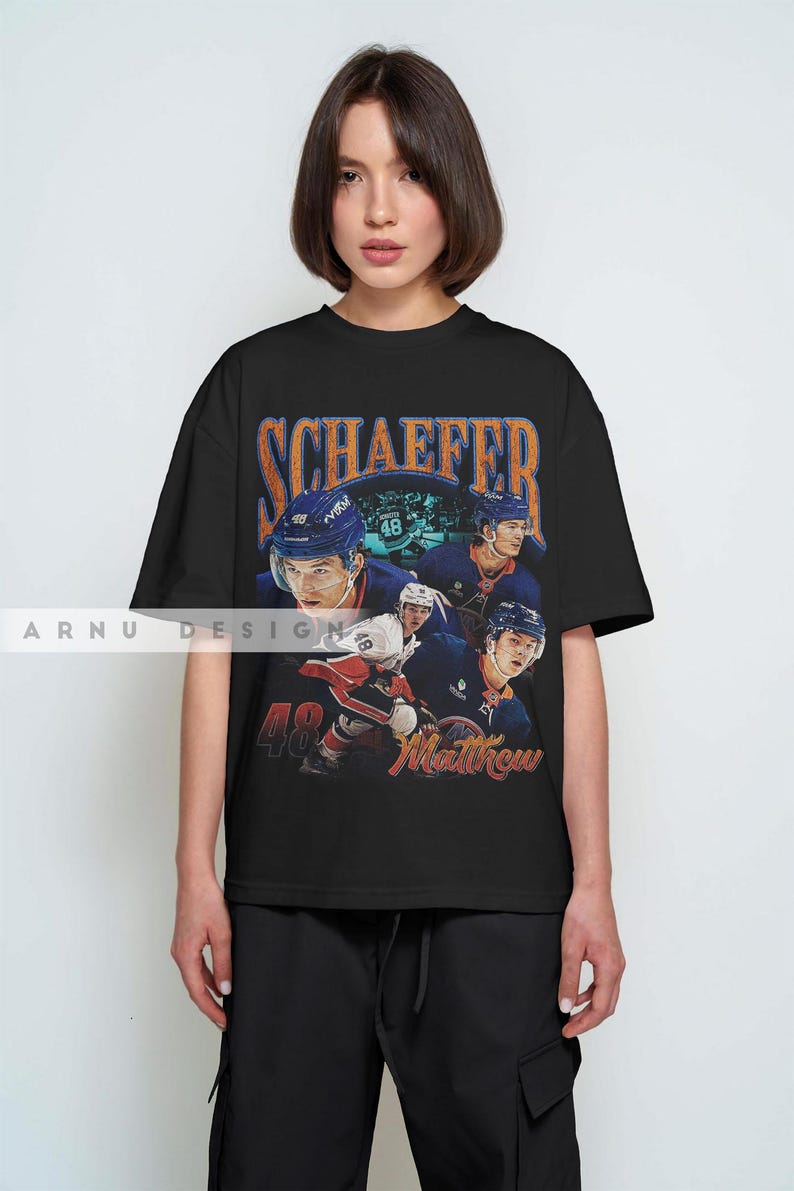 May include: Black t-shirt featuring a vintage-style graphic of a hockey player in a blue uniform. The design includes the name "SCHAEFER" in orange lettering and the number "48" with the name "Matthew". The shirt is a casual wear item.