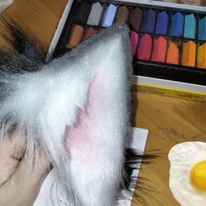 May include: A fluffy, white and black faux fur cat ear with a pink interior. The ear is held up, with a box of colorful chalk pastels in the background. The ear has a glittery texture.