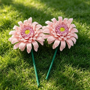 Könnte beinhalten: Zwei handgefertigte Häkelblumen mit hellrosa Blütenblättern, einer strukturierten rosa Mitte und einem leuchtend gelben Kern. Jede Blume ist an einem dünnen, geraden grünen Stiel befestigt, was eine detaillierte botanische Darstellung aus Garn ergibt.