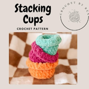 May include: A stack of four crocheted cups in different colors: orange, pink, teal, and green. The cups are stacked on a black and white checkered surface. The text "Stacking Cups CROCHET PATTERN" is above the cups.