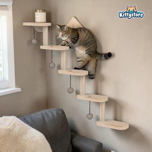 May include: A wooden cat climbing shelf system with multiple platforms and a triangular top piece. A tabby cat is climbing the structure. Each platform has a hanging ball toy. The Kittystore logo is visible in the top right corner.