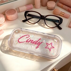 May include: A clear rectangular case with the name "Cindy" spelled out in pink rhinestones, along with a pink star. Black-framed eyeglasses and various pink makeup items are arranged on a white surface.