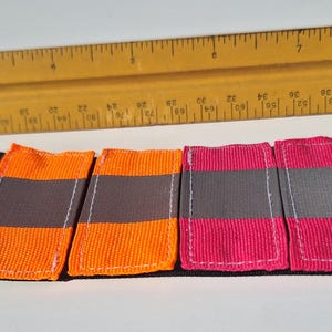 May include: Four colorful fabric patches with a gray stripe, orange and pink. A wooden ruler is in the background. The patches are on a black fabric base.