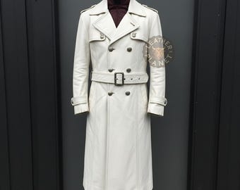 May include: A white leather trench coat with a double-breasted design, featuring a belt at the waist and button details. The coat has a classic collar and epaulettes on the shoulders. The coat is displayed on a mannequin.