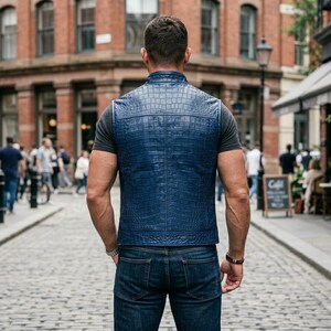 May include: A person wearing a blue, crocodile-patterned vest and dark blue jeans stands on a cobbled street. The vest is the main focus, with a textured finish. The scene is set in a city with brick buildings.