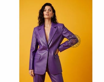 Può includere: Un vivace completo composto da blazer e pantaloni viola. Il blazer presenta un classico risvolto a lancia, una chiusura a un bottone e due tasche anteriori. L'outfit è realizzato in un materiale liscio simile alla pelle, creando un look elegante e moderno. Lo sfondo è giallo.