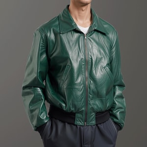 May include: A green leather bomber jacket with a collared neckline and a zip-up front. The jacket features two front pockets and a black ribbed hem. The sleeves are long, and the jacket is worn over a white t-shirt and dark blue pants.