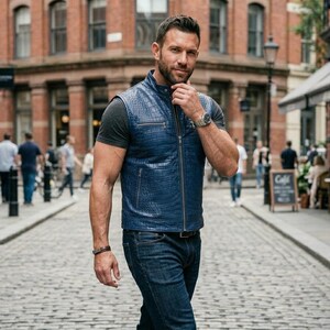 May include: A man in a blue leather vest with a textured design, a grey t-shirt, and blue jeans. He wears a watch and bracelet. The background is a cobbled street with brick buildings.