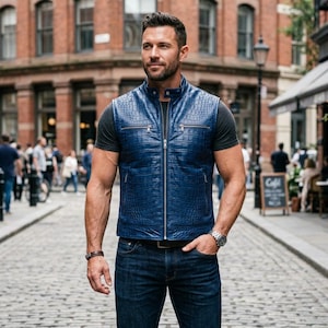 May include: A blue leather vest with a crocodile pattern, featuring a zip-up front and a stand-up collar. The vest is worn over a grey t-shirt and paired with blue jeans. A silver watch and a bracelet are visible.