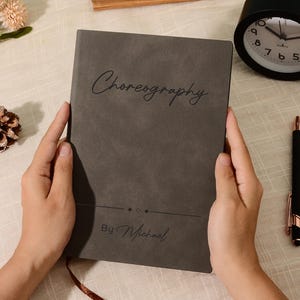 Könnte beinhalten: Ein graues Notizbuch mit dem Wort "Choreography" in Schreibschrift. Der Text "By Michael" steht unter dem Titel. Ein schwarzer Stift mit goldenen Akzenten und ein schwarzer Wecker sind ebenfalls zu sehen.
